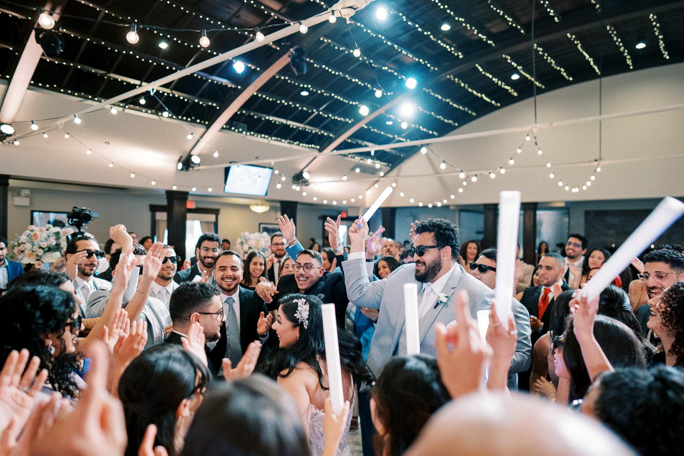 A lively group of wedding guests celebrating at a reception under string lights, with people smiling, dancing, and holding glow sticks in a decorated venue.