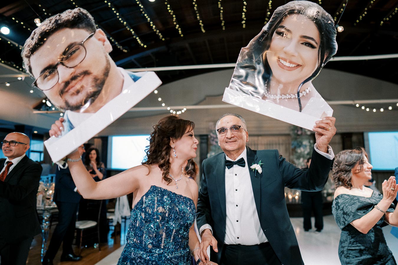 A joyful wedding reception scene with elegantly dressed guests holding large cut-out photos of a smiling couple. The venue is decorated with string lights, creating a festive atmosphere.
