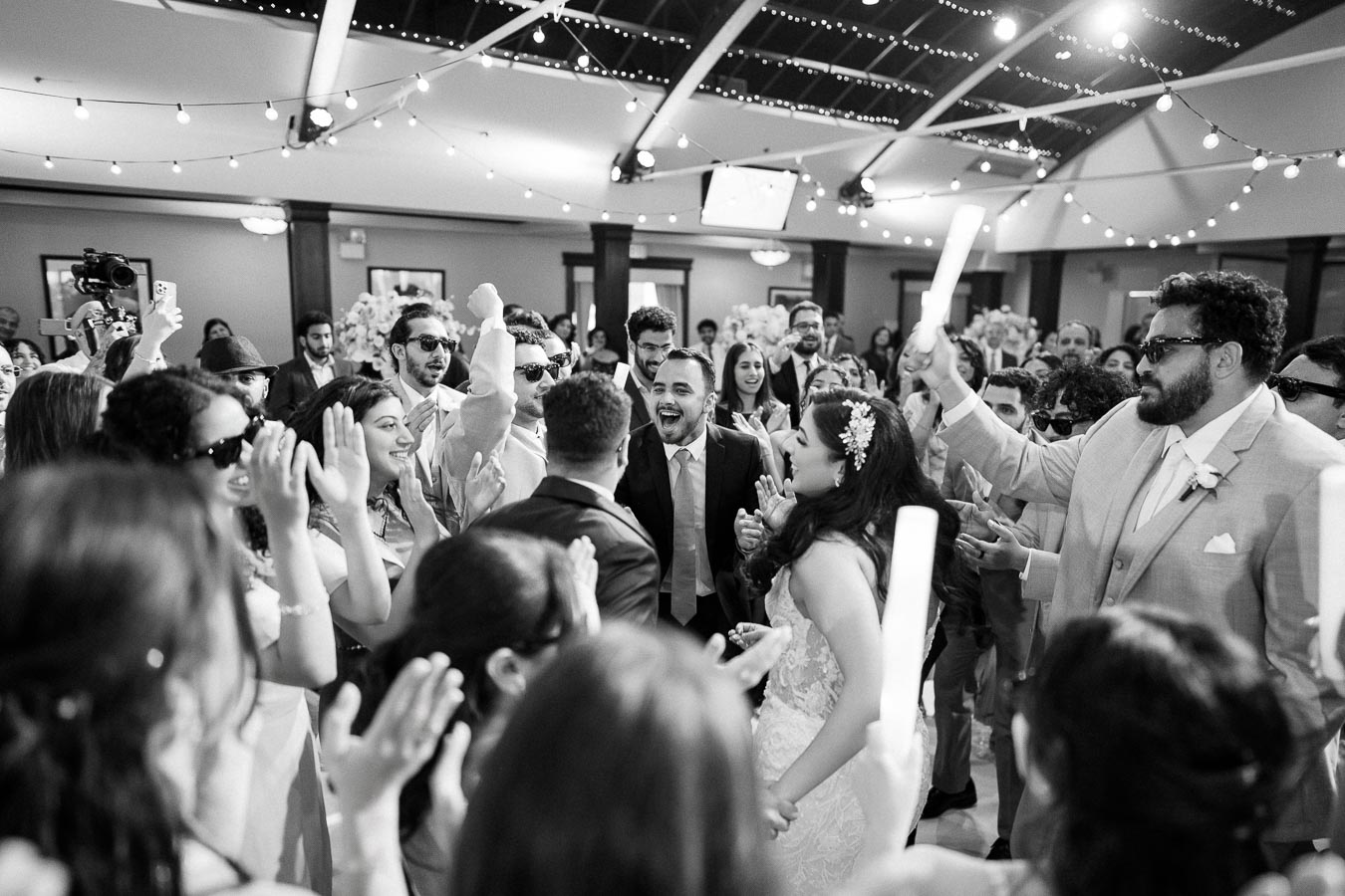 A joyful wedding celebration with a bride and groom dancing surrounded by cheering guests, under string lights in an elegant reception venue.