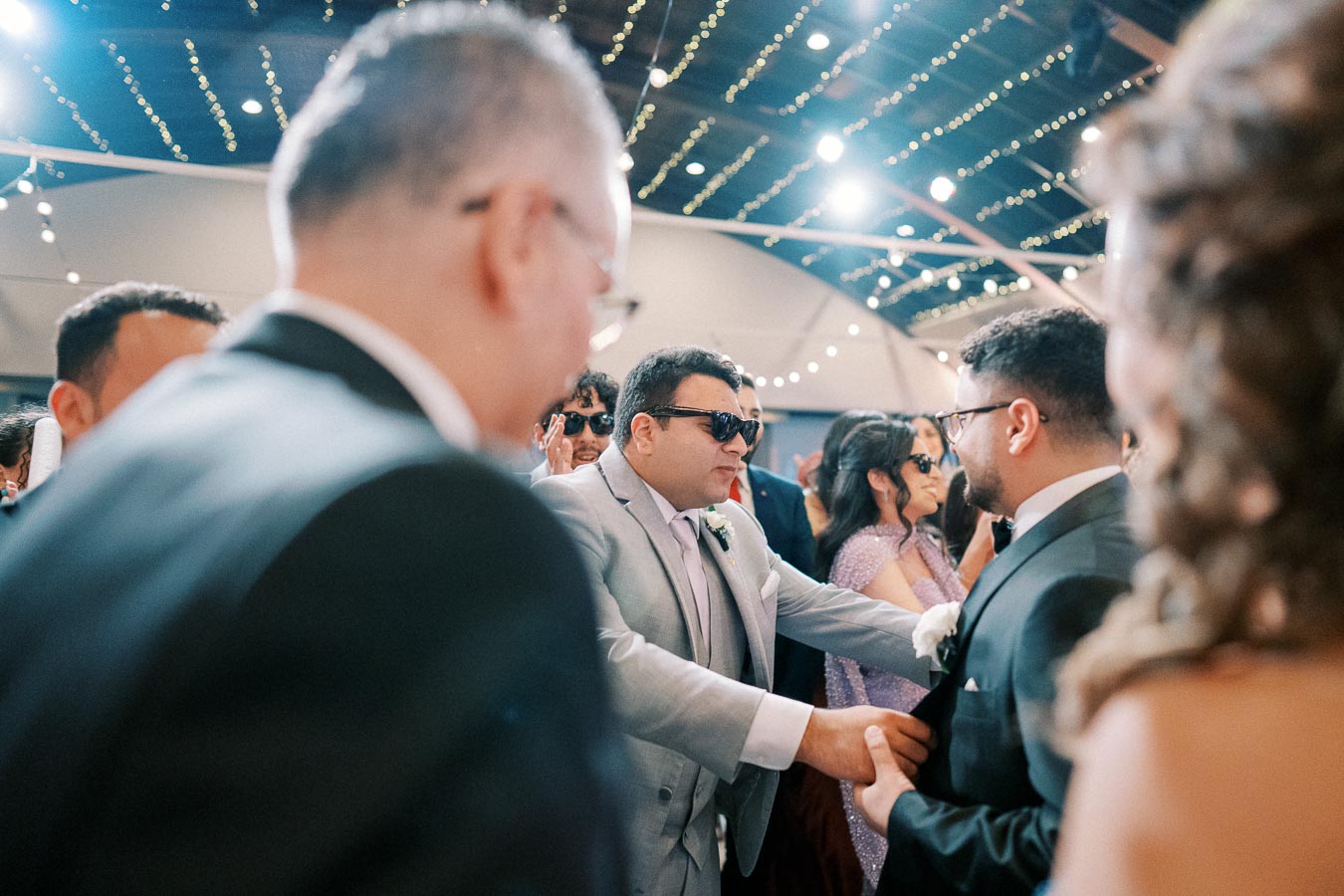 A group of people celebrating at a wedding reception, with men in suits and women in formal dresses under string lights.