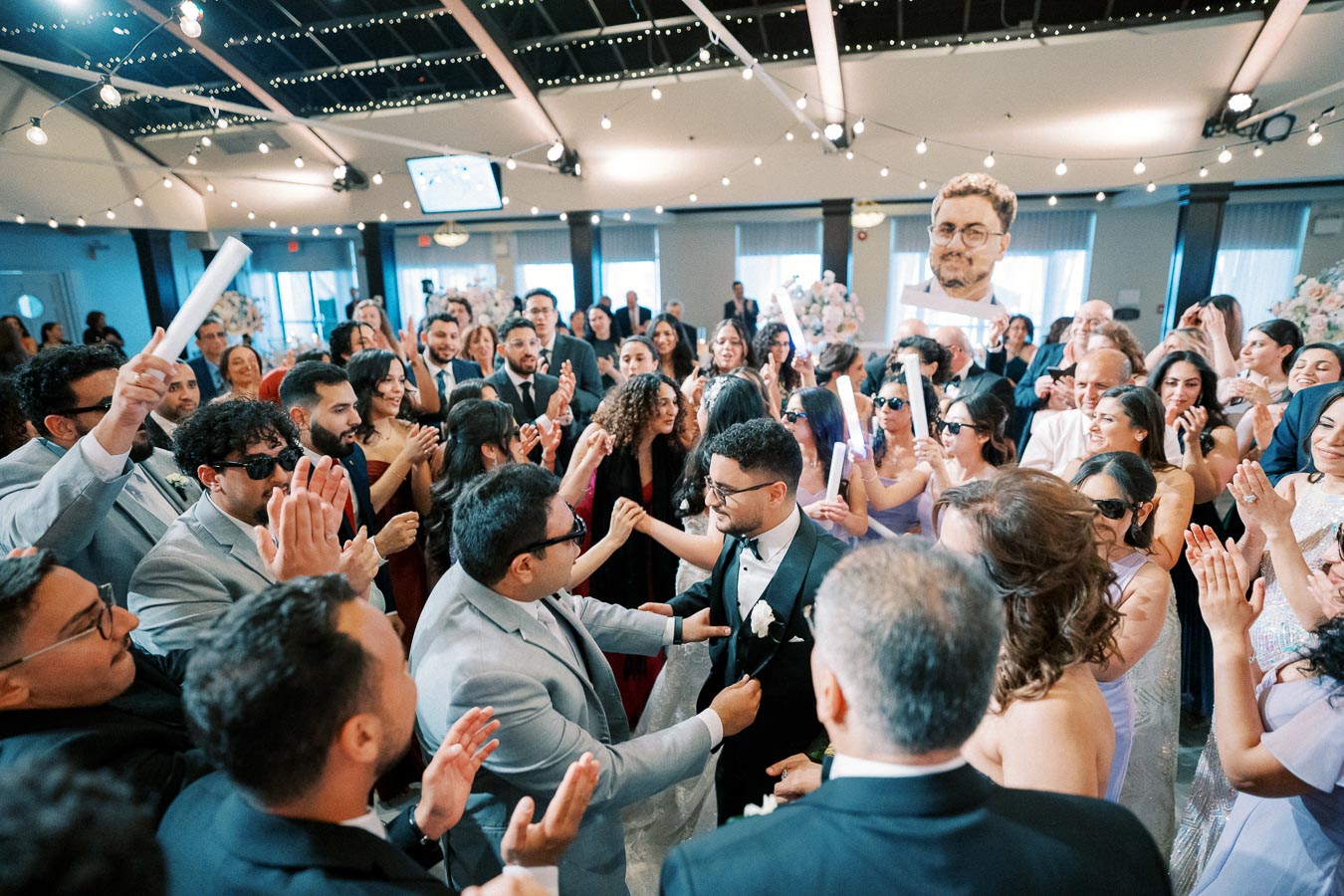 Wedding celebration with a groom surrounded by joyful guests, dancing and clapping in a beautifully decorated venue with string lights and elegant attire.