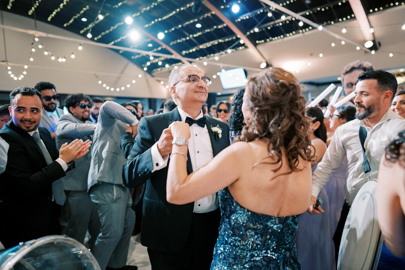 Group enjoying a lively celebration on a dance floor, under string lights in a wedding or party setting, with a couple dancing at the center while others applaud and join the festivities.