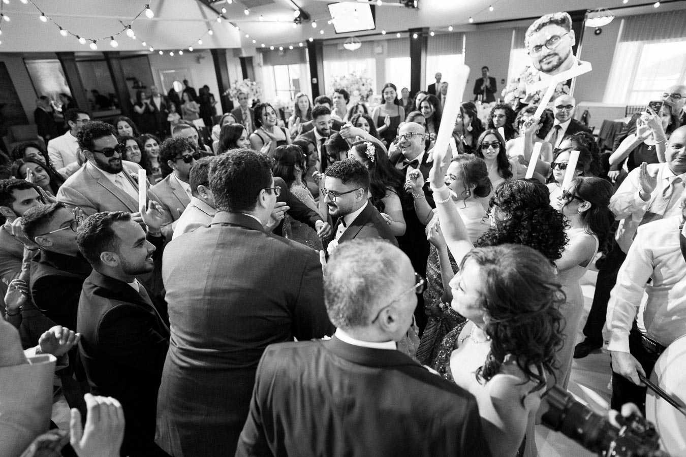 A lively black and white image of a wedding reception with a large group of guests joyfully dancing and celebrating. The festive atmosphere includes people holding foam sticks and a humorous cutout, all under string lights in a decorated venue.