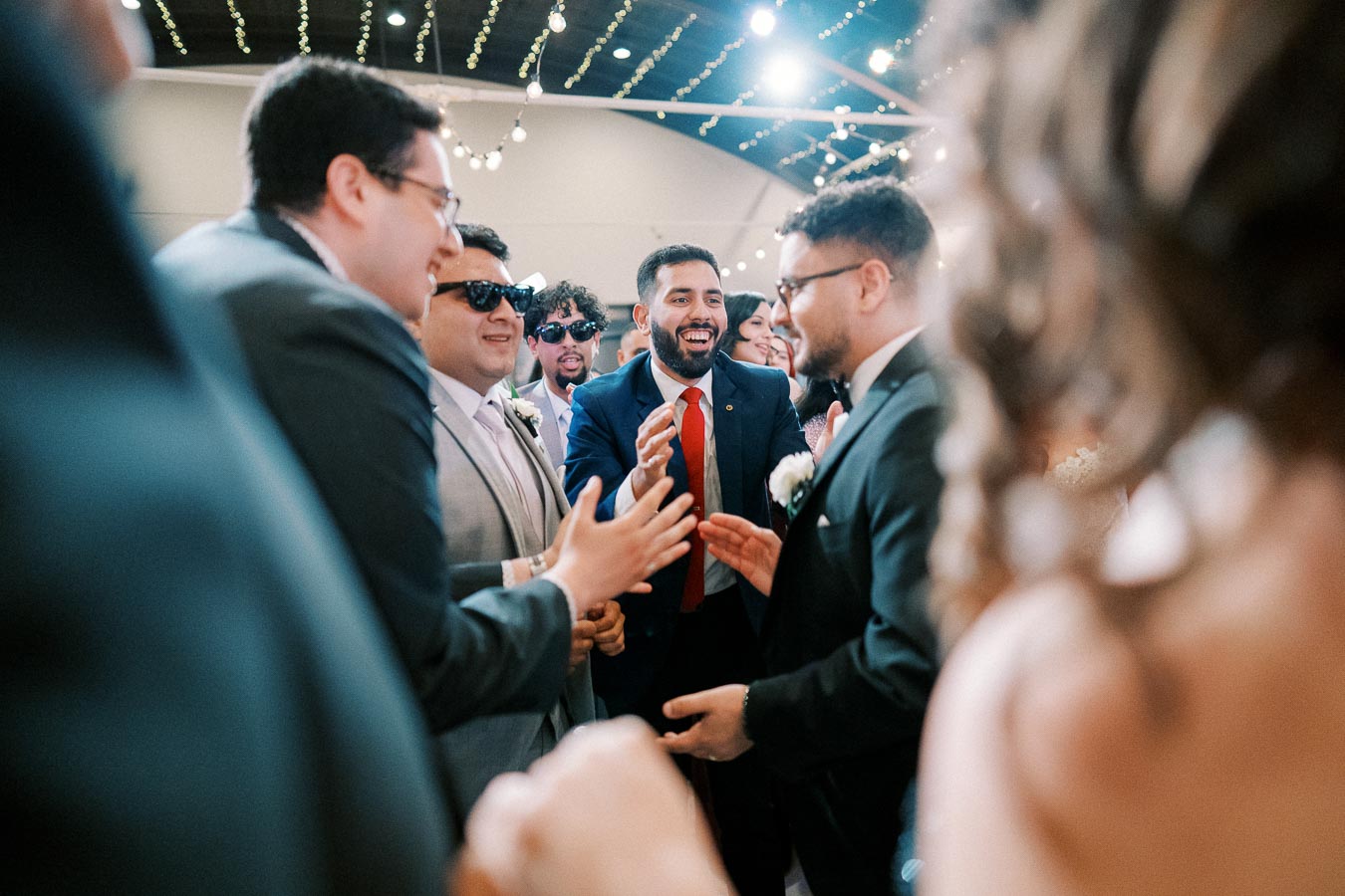 A group of well-dressed men, smiling and engaging in lively conversation at a wedding reception, under decorative string lights.