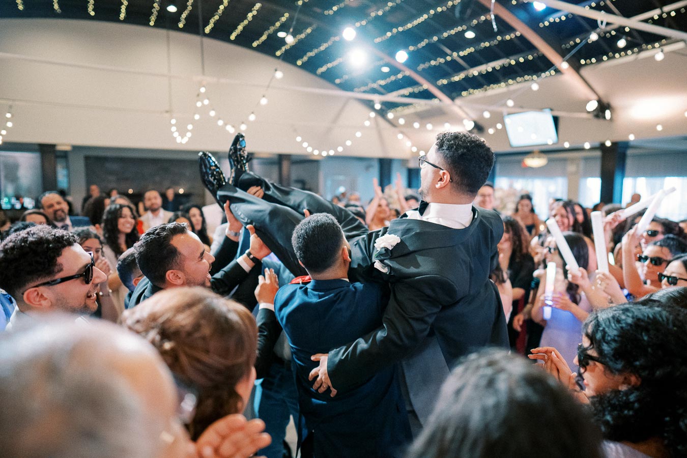 A lively wedding reception scene with a groom being lifted by friends amidst a cheering crowd, adorned with twinkling string lights and joyous guests celebrating together.