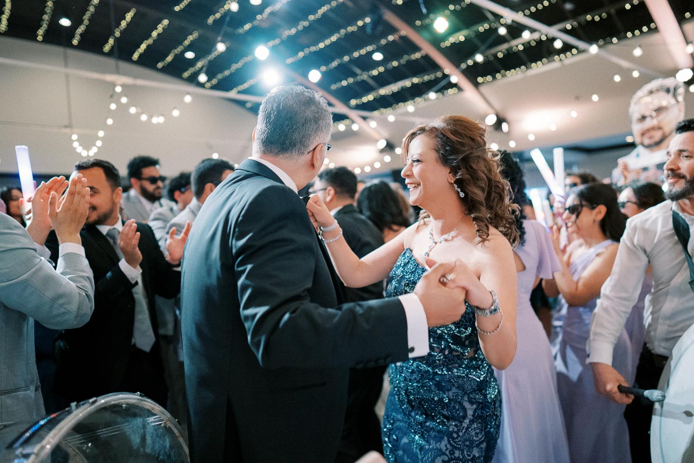 A couple dancing joyfully at a lively wedding reception, surrounded by cheering guests and sparkling string lights.