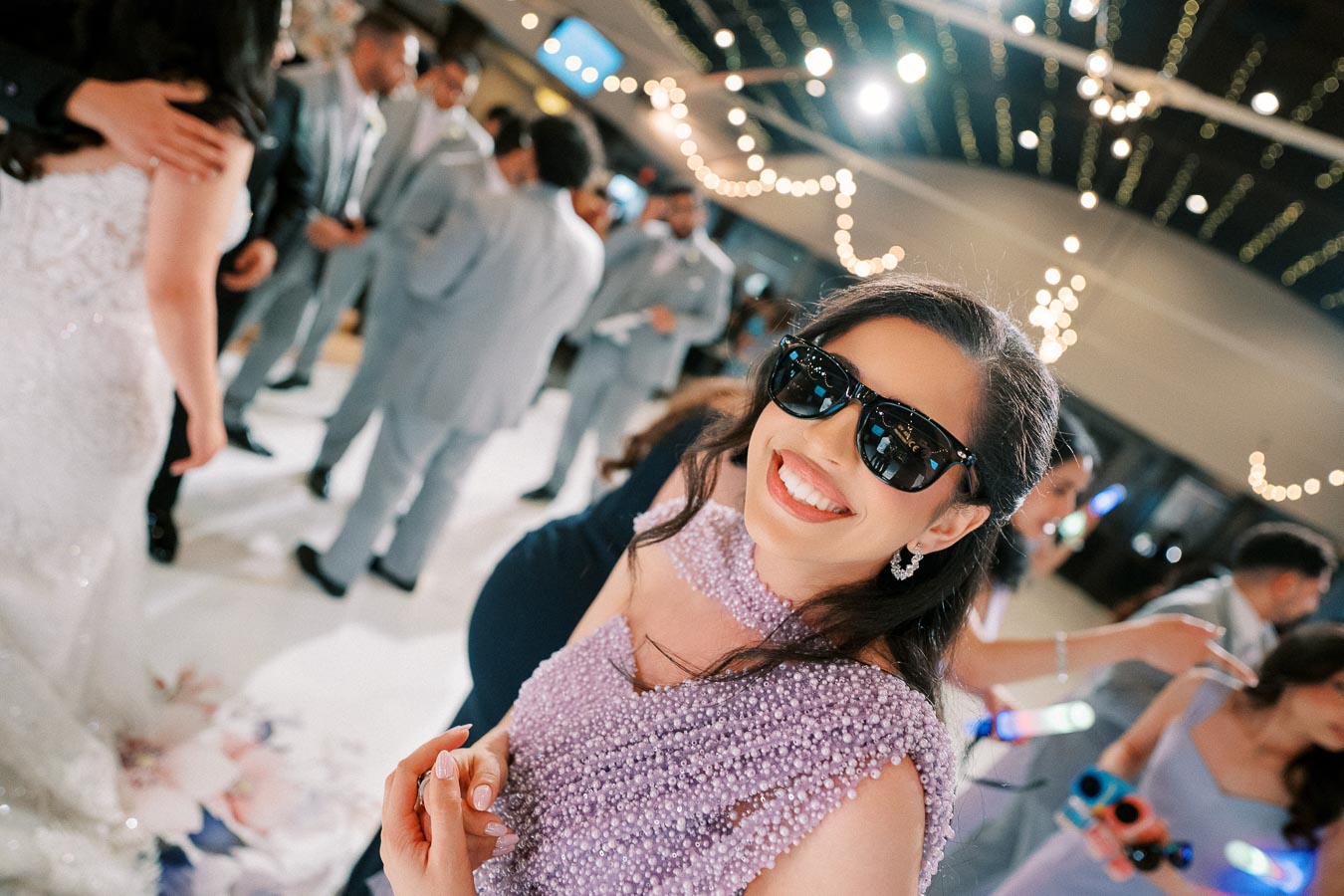 Smiling woman in a beaded lavender dress and sunglasses at a lively wedding reception, with guests in formal attire and string lights in the background.