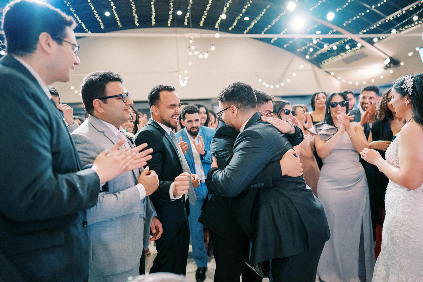 A joyful wedding reception with a group of well-dressed guests, including men in suits and women in dresses, celebrating and clapping as two men embrace. The festive atmosphere includes string lights and smiling faces, capturing a moment of friendship and celebration.