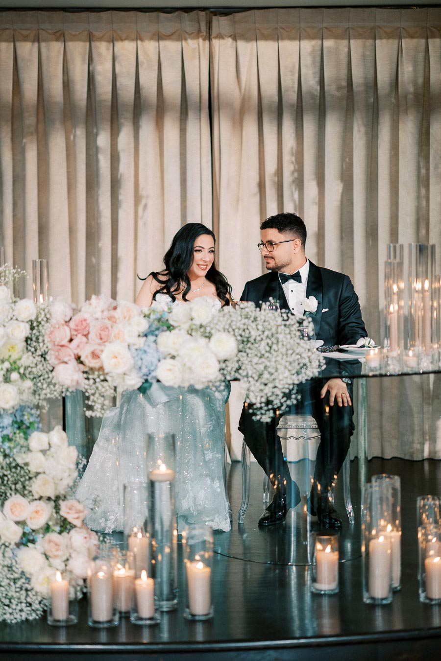 Elegant wedding scene with a bride in a shimmering gown and a groom in a black tuxedo sitting at a floral-adorned table, surrounded by candles and soft lighting.