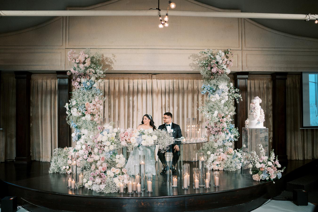 Elegant wedding reception setup with a bride and groom seated at a floral-adorned table. The scene is decorated with tall, colorful floral arrangements and glowing candles, with a tiered wedding cake displayed on a stand. The backdrop is luxurious with dark wood and soft curtains, creating a romantic and sophisticated ambiance.