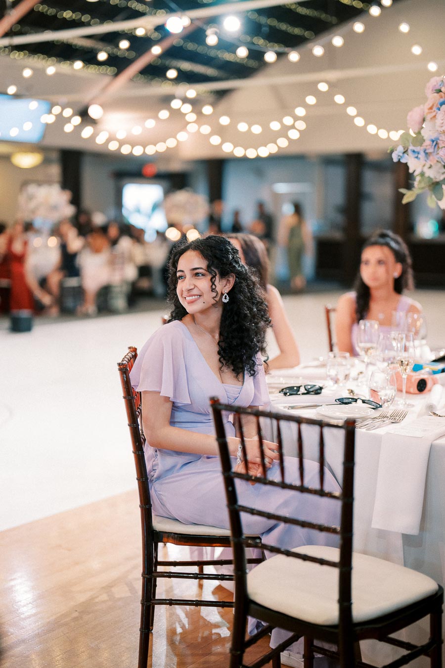 A woman in a light purple dress sits at a beautifully decorated table with elegant glassware and floral centerpieces at a wedding reception, surrounded by warm string lights and a lively atmosphere.