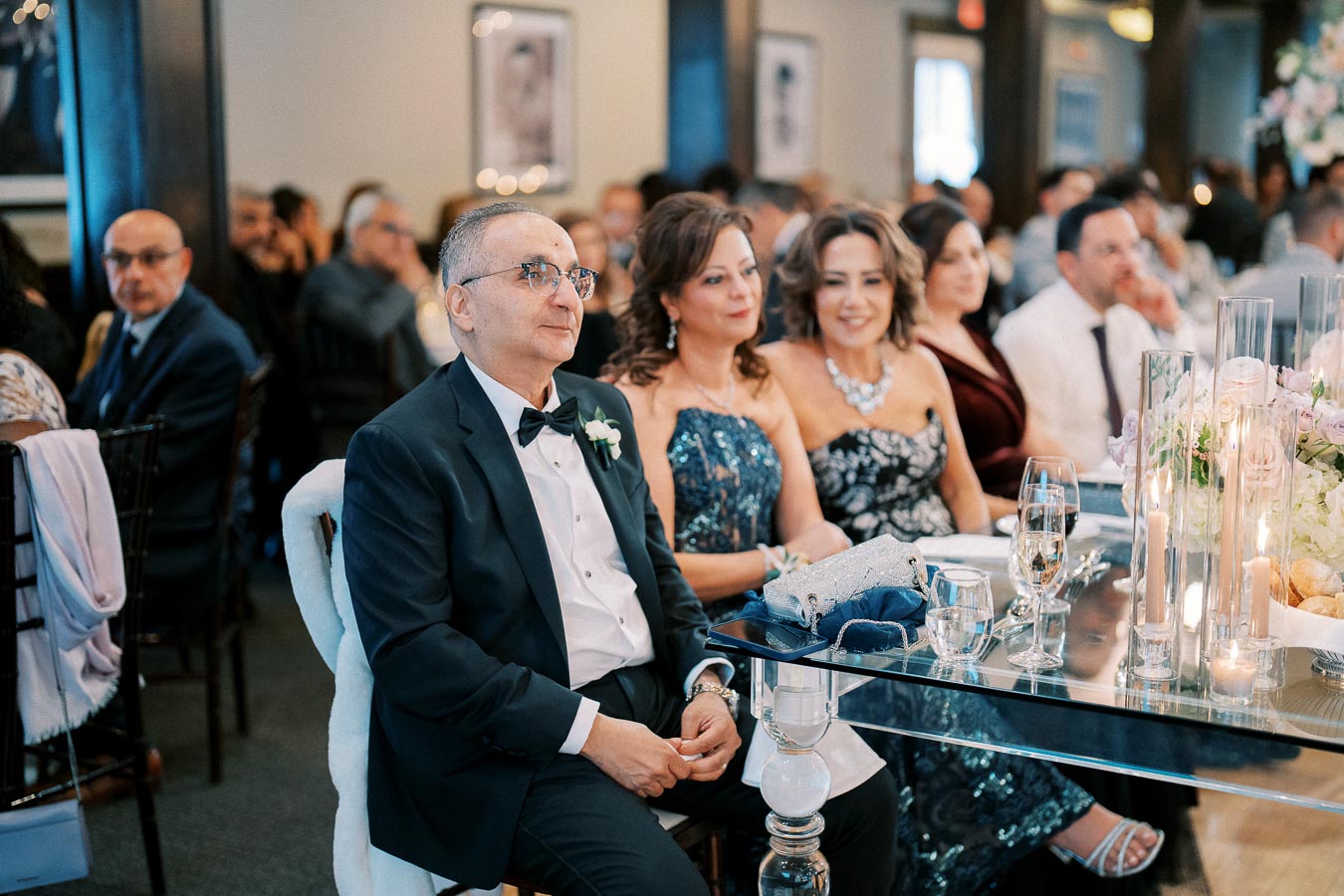 A group of elegantly dressed guests sitting at a wedding reception, featuring a man in a tuxedo and women in formal gowns, with flowers and candles decorating the table in the foreground.