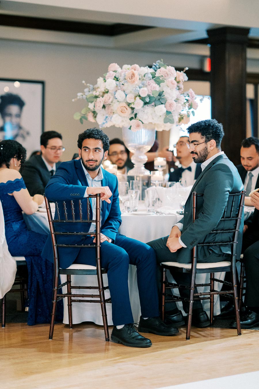 A group of elegantly dressed people sit at a round table adorned with a large floral centerpiece during a formal event.