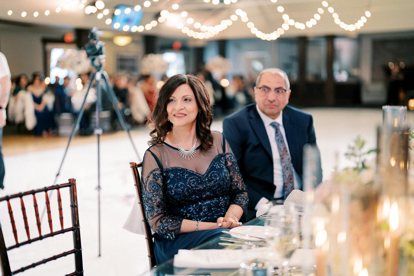 Elegant couple seated at a formal event, surrounded by a beautifully decorated venue with string lights and a lively atmosphere in the background.
