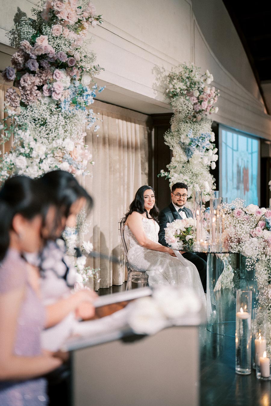Elegant wedding reception with a couple seated amidst beautiful floral decorations and candles, as a guest gives a speech.