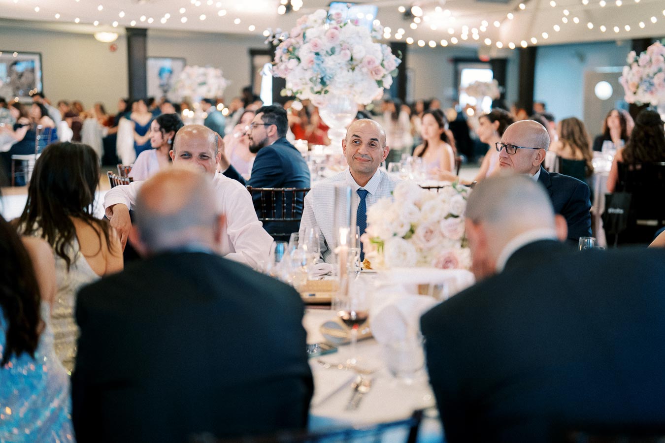 Elegant wedding reception with guests seated around a beautifully decorated table, featuring floral centerpieces and soft ambient lighting.