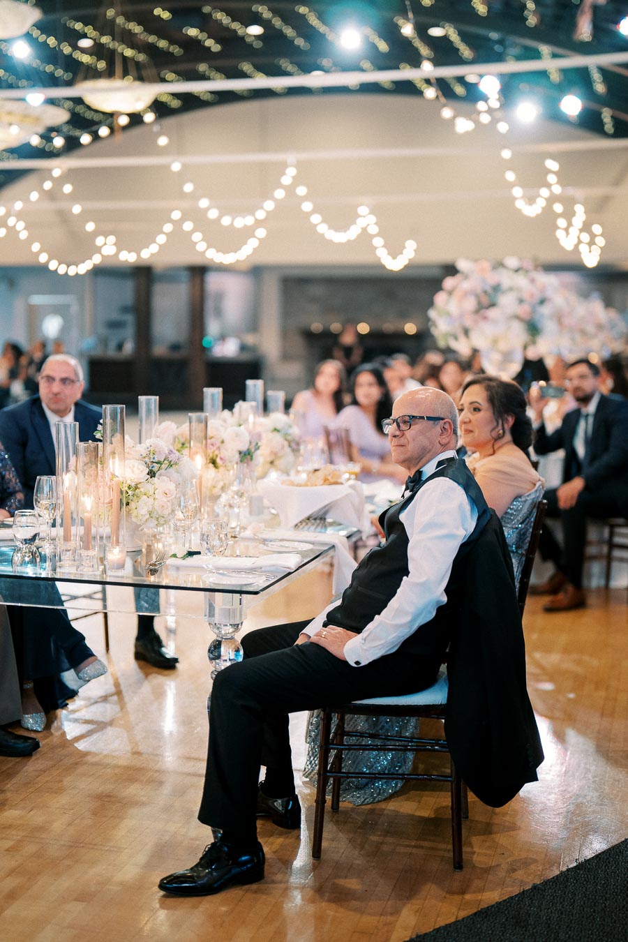 Elegant wedding reception with guests seated at a beautifully decorated table, adorned with white floral arrangements and candles, under romantic string lights.