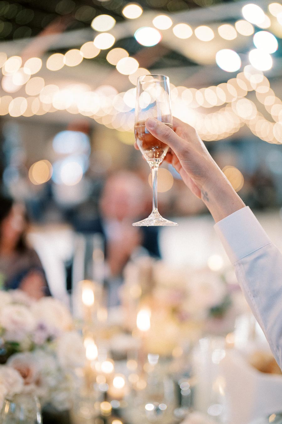 A celebratory setting with a hand holding a champagne flute against a backdrop of soft, glowing string lights and blurred guests, creating a festive atmosphere.