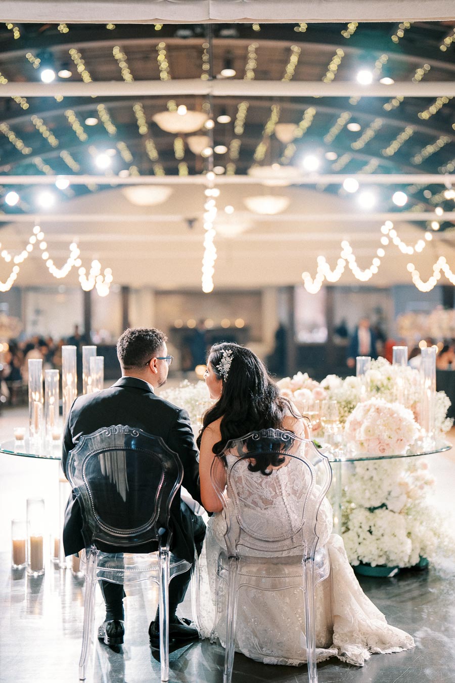 Elegant wedding reception with a bride and groom sitting on transparent chairs at a beautifully decorated table, adorned with white and blush flowers under twinkling string lights.