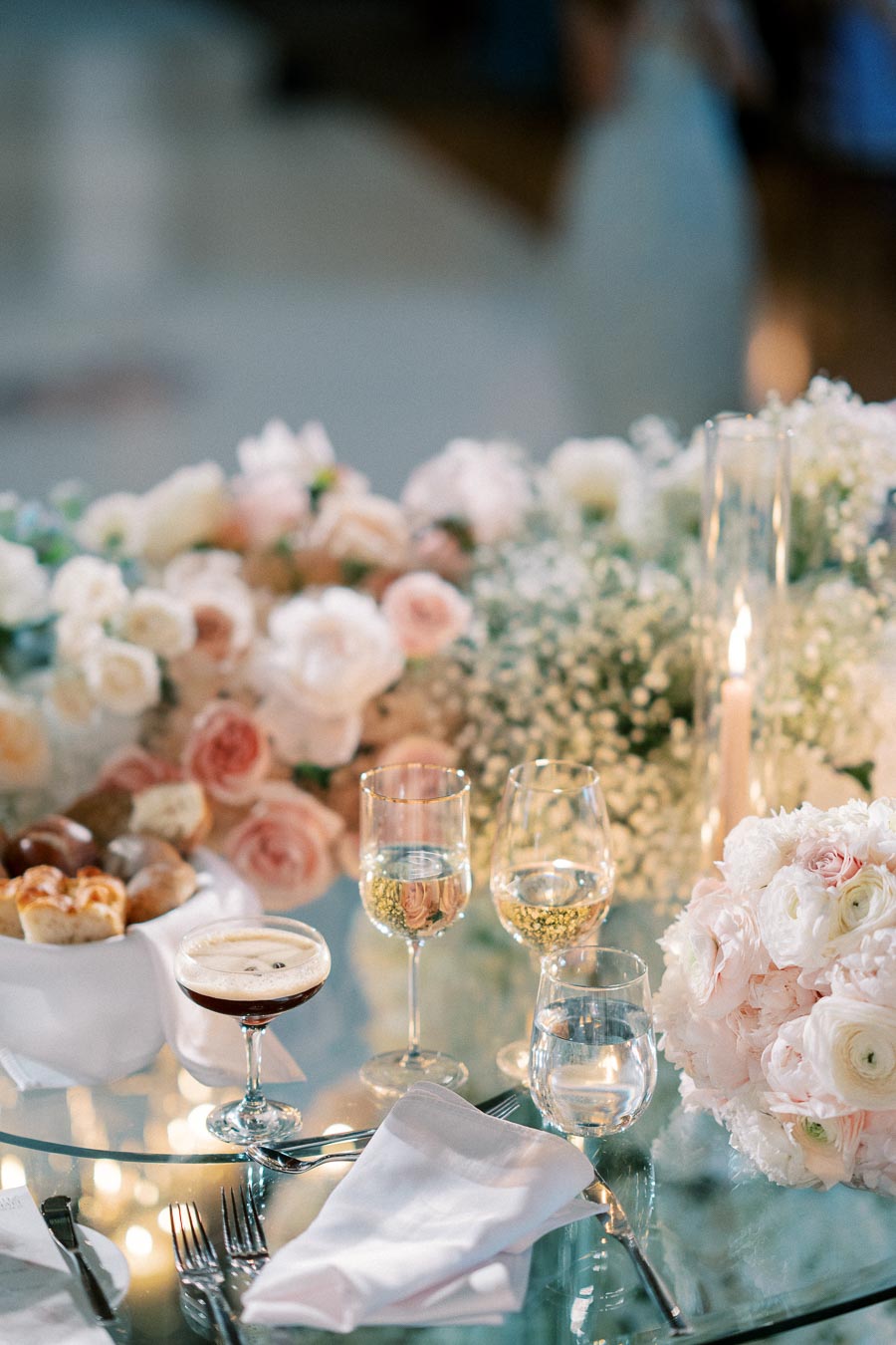 Elegant wedding table setting with white and pink floral arrangement, wine glasses, and candles on a reflective glass surface.