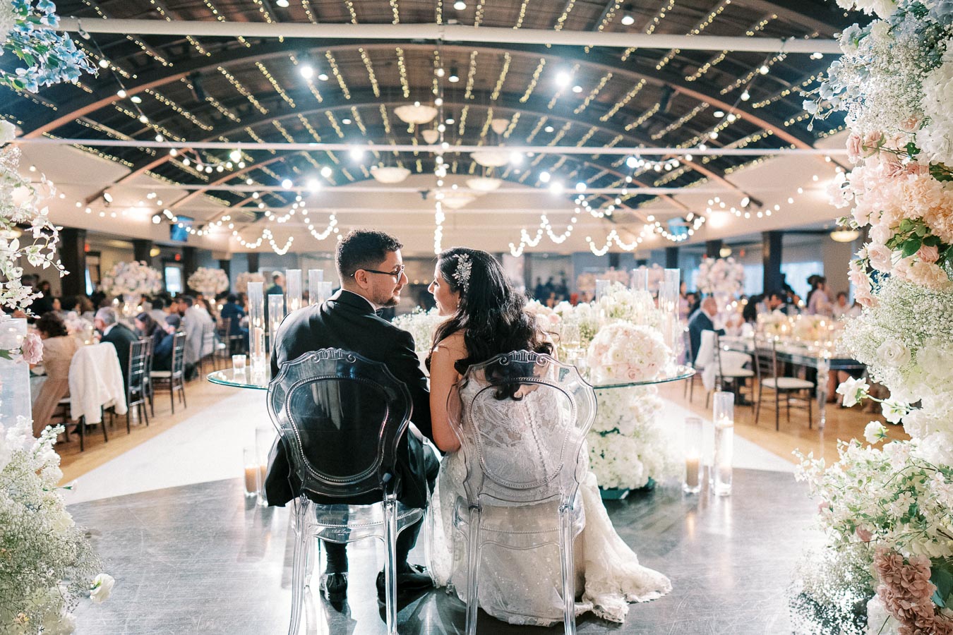 A newlywed couple sits at a beautifully decorated wedding reception venue, surrounded by elegant floral arrangements and twinkling string lights, creating a romantic atmosphere.