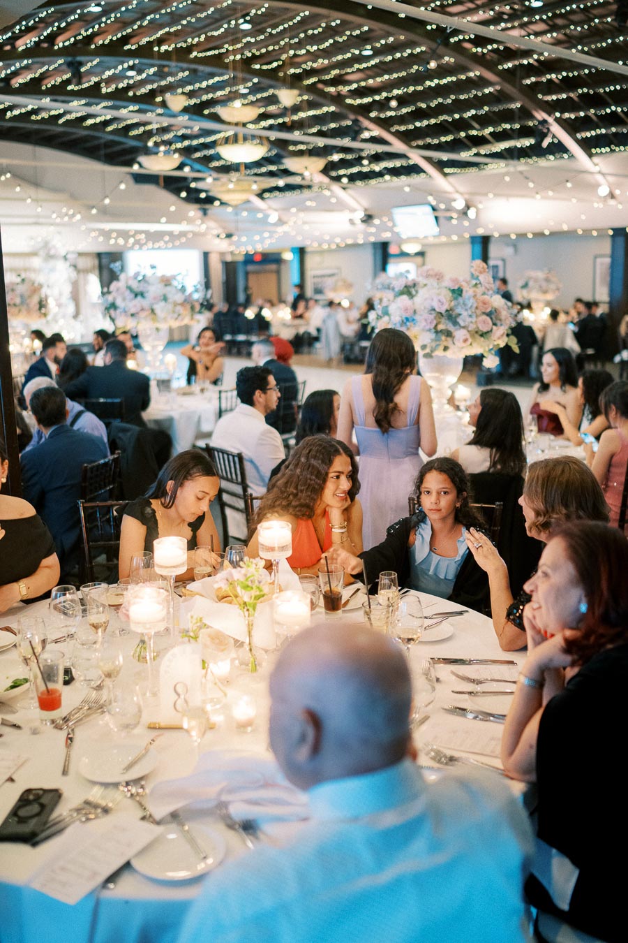 A vibrant and elegant indoor wedding reception with guests seated around tables adorned with candles and floral centerpieces, under a ceiling decorated with string lights, creating a festive and warm atmosphere.