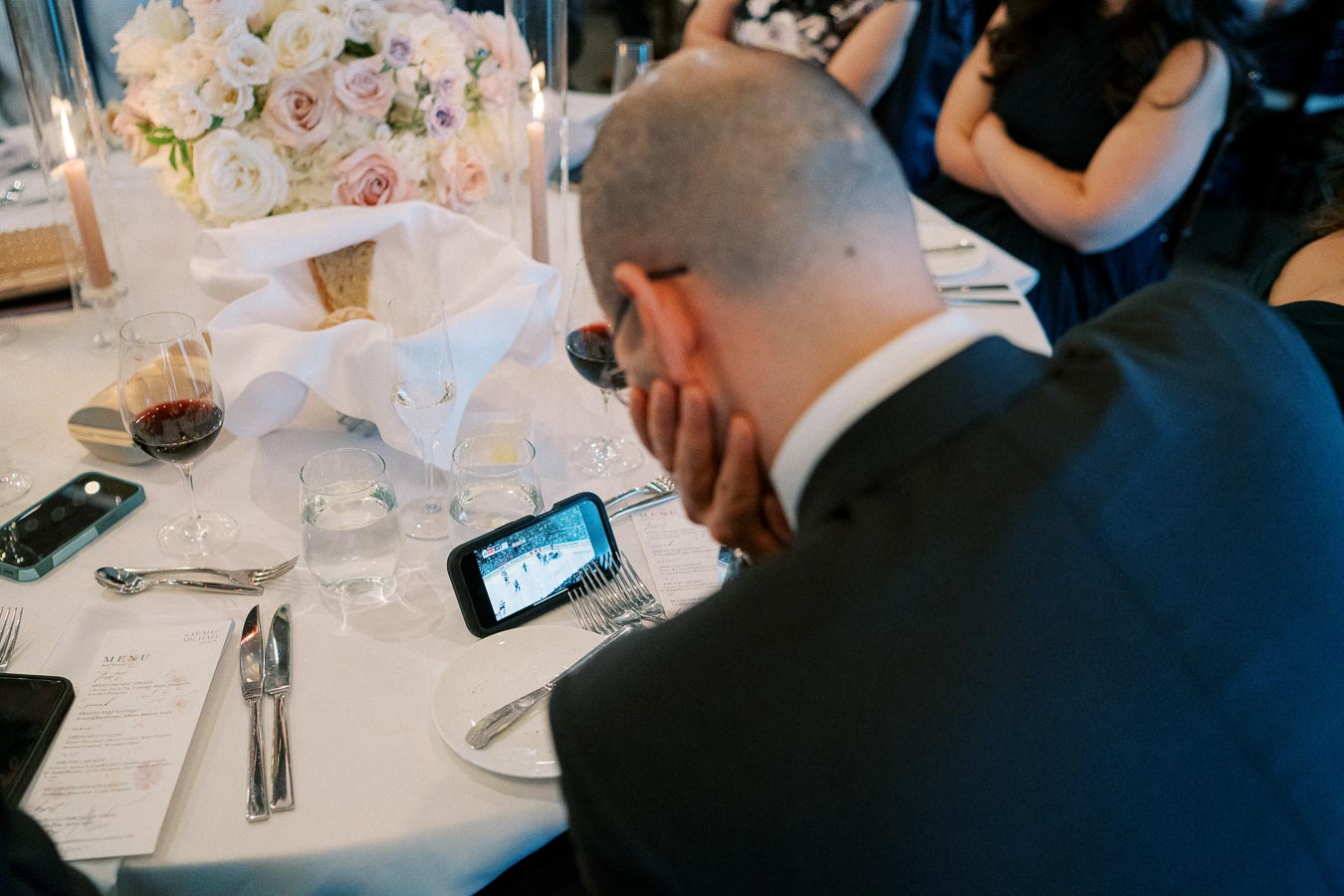 Man in a suit watching a video on a smartphone at a banquet table set with wine glasses, utensils, a menu, and a floral centerpiece.