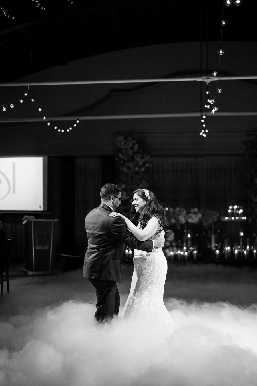 A bride and groom share their first dance on a misty dance floor, surrounded by soft lighting and romantic decorations.