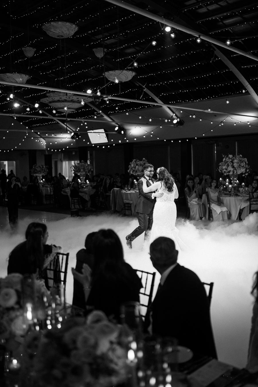 Newlywed couple dancing at their wedding reception under twinkling string lights and surrounded by seated guests, with a dreamy cloud-like effect on the dance floor in a beautifully decorated venue.