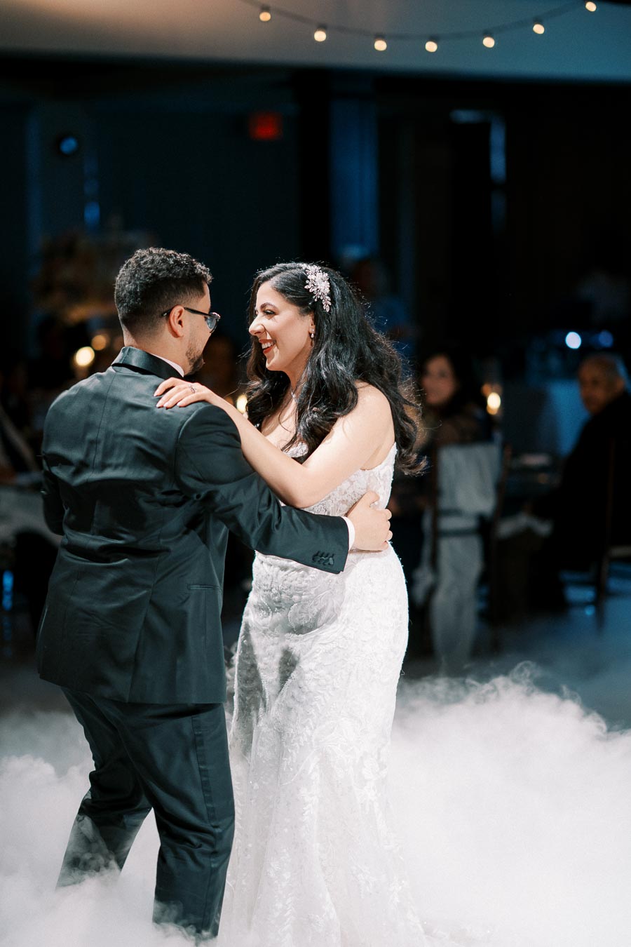 A bride and groom share their first dance on a cloud-filled dance floor at a wedding reception, surrounded by soft, romantic lighting and guests in the background.