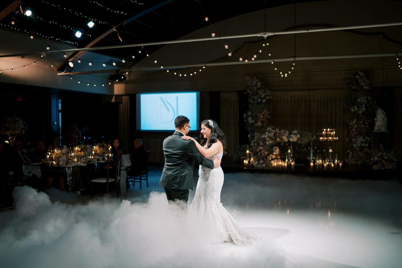 A bride and groom share their first dance at a beautifully decorated wedding reception, surrounded by mist and fairy lights. The elegant venue features a floral backdrop and warm candlelight, creating a romantic atmosphere.