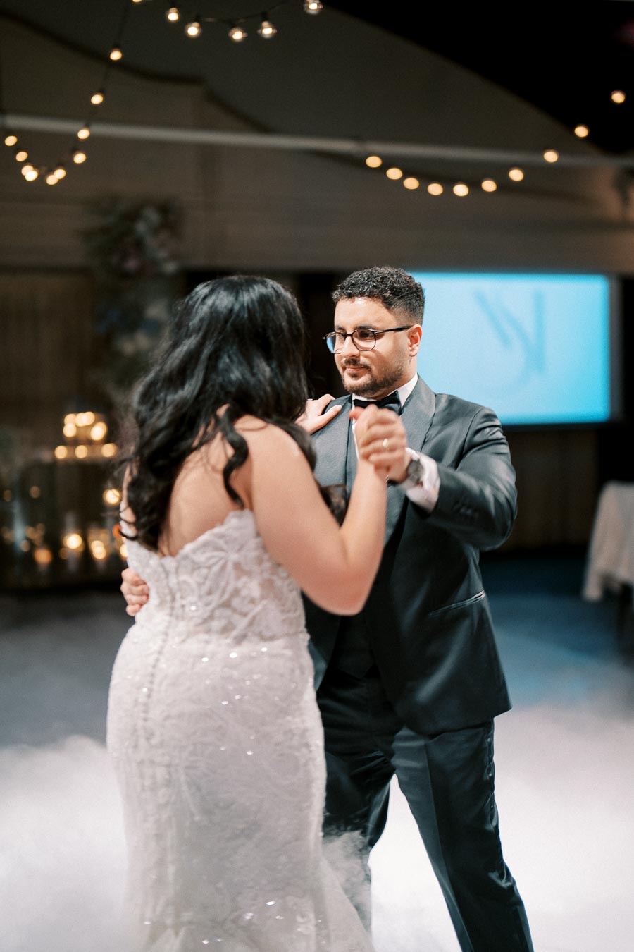 A couple elegantly dancing at a wedding reception, with ambient string lights in the background, capturing a romantic moment on the dance floor.