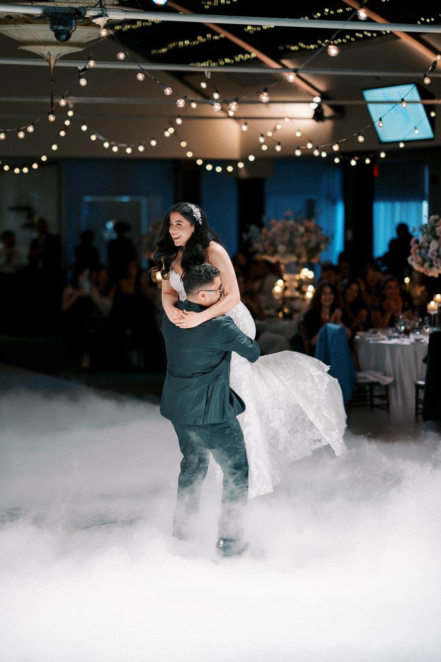 A groom joyfully lifts his bride during their first dance at a wedding reception, surrounded by soft clouds of smoke and elegant string lights.