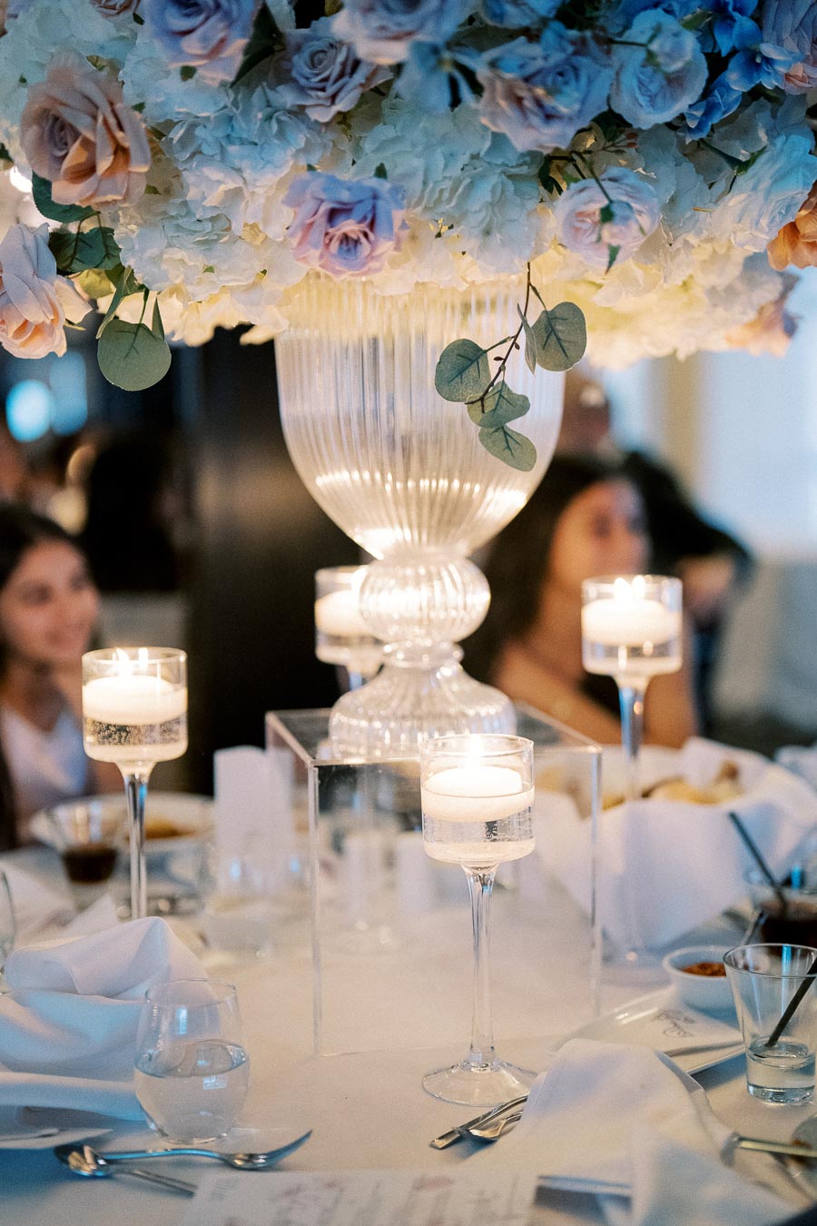 Elegant wedding reception table setting featuring a large floral centerpiece with pastel roses and hydrangeas, surrounded by floating candle holders on a white tablecloth.