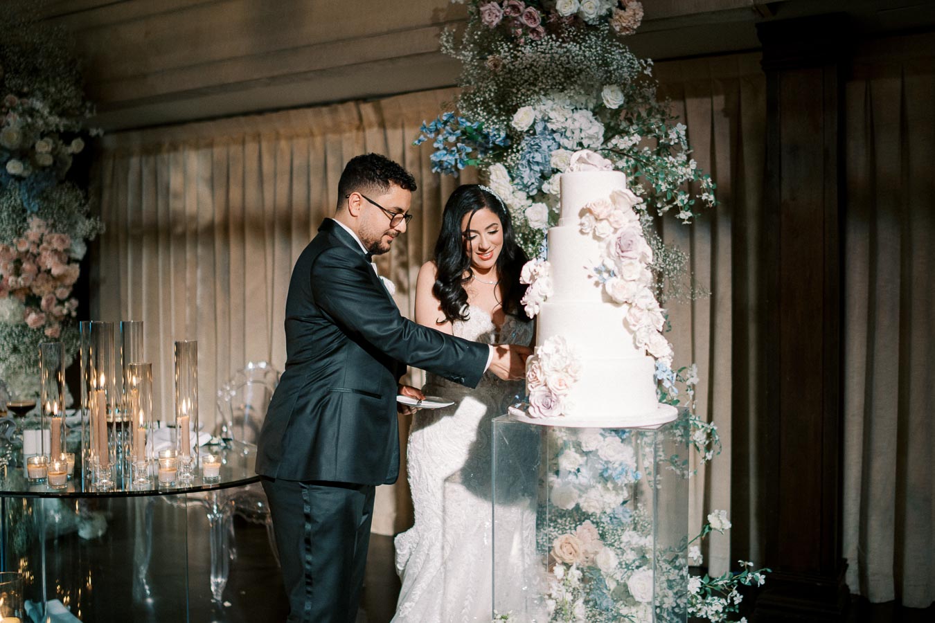 A bride and groom cutting an elegant white tiered wedding cake adorned with pastel flowers, surrounded by candles and floral decorations, in a warmly lit reception venue.