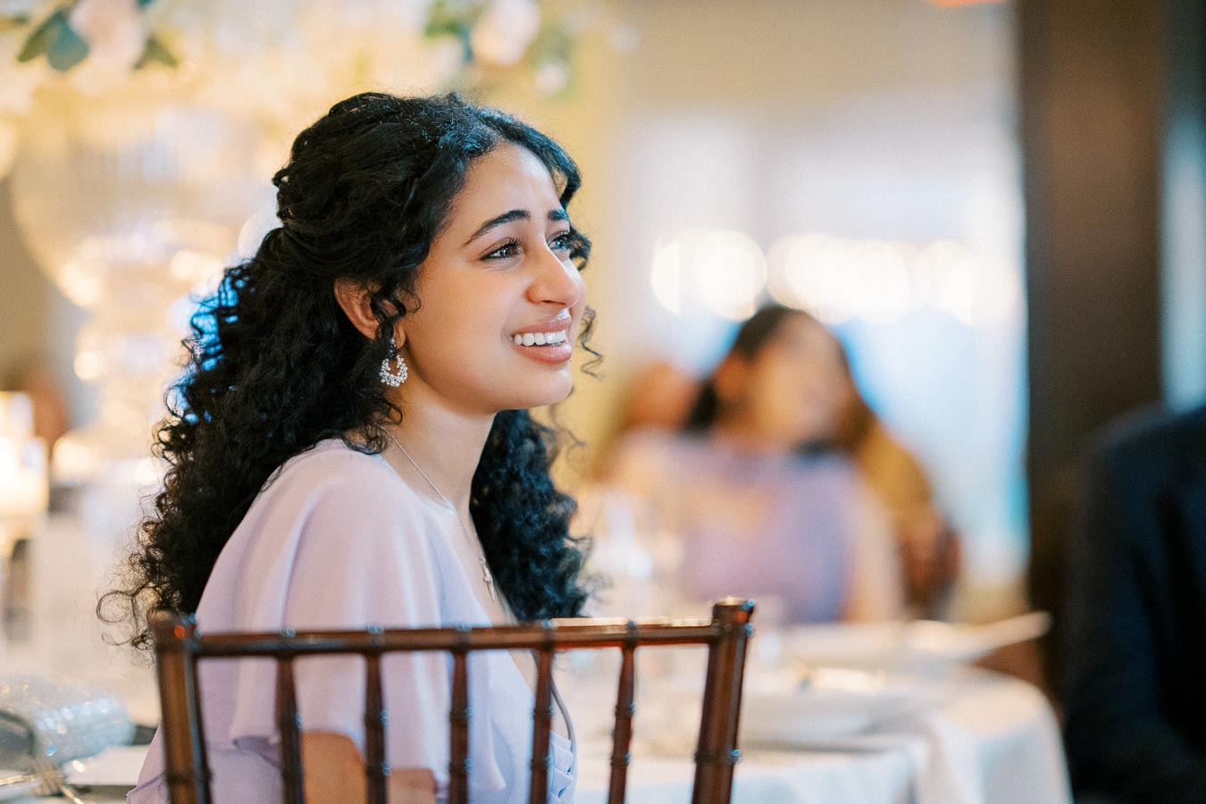 A woman with curly hair and elegant earrings smiles while seated at a formal event, surrounded by a softly blurred background.