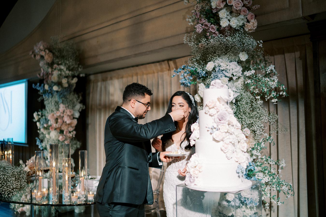 A bride and groom share a moment during their wedding reception as the groom feeds the bride a piece of cake. The scene is elegantly decorated with a multi-tiered white wedding cake adorned with pastel flowers, set against floral arrangements and gentle candlelight.