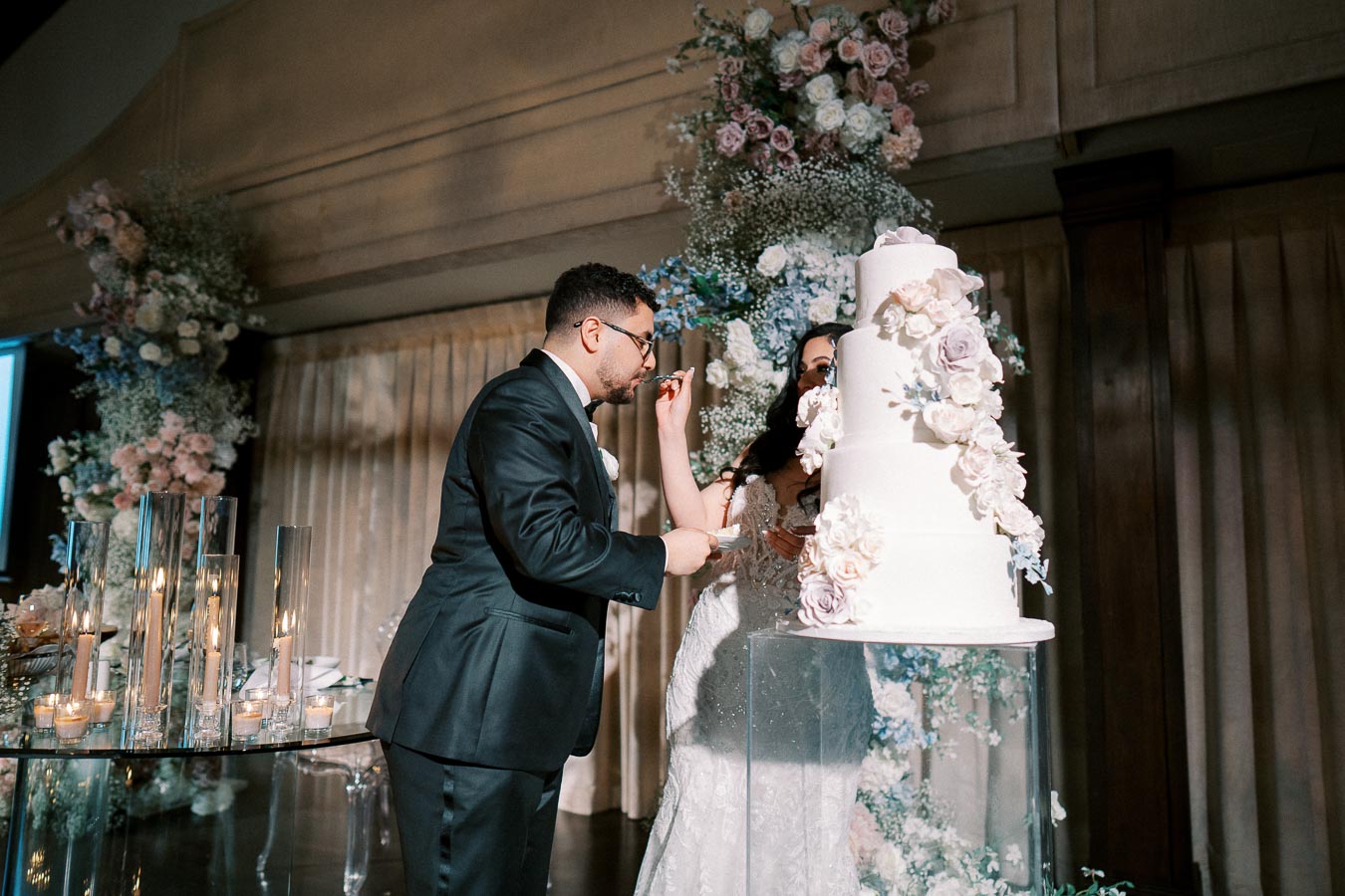 A bride and groom stand by a beautifully decorated, multi-tiered wedding cake adorned with floral accents. The groom, dressed in a black tuxedo, is being fed a slice of cake by the bride, who wears an elegant white dress. The background is filled with an arrangement of flowers and soft lighting, creating a romantic atmosphere. Candles in glass