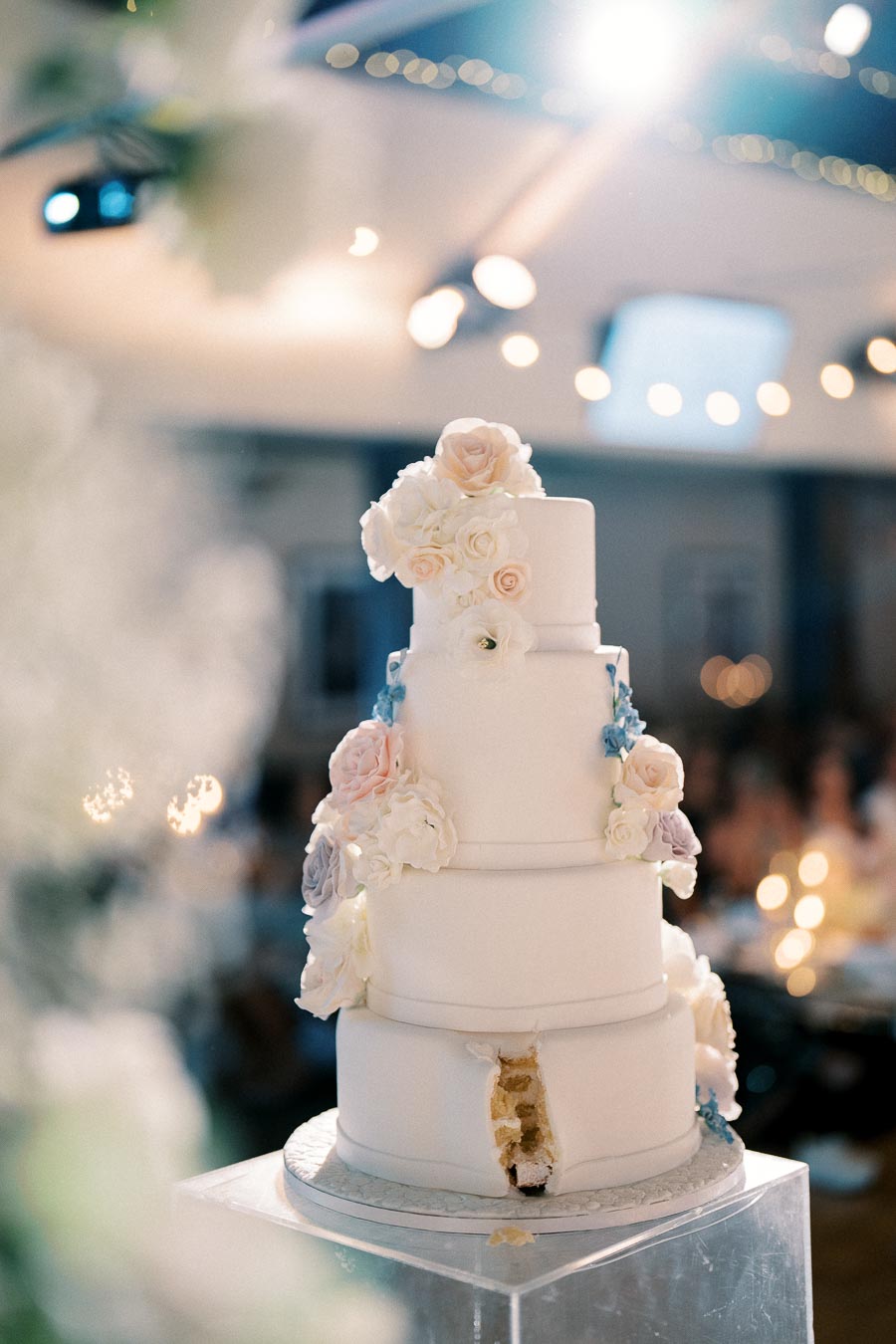 Four-tier white wedding cake adorned with delicate pastel flowers, showcasing intricate floral designs and a small cut section, set in an elegantly lit reception venue.