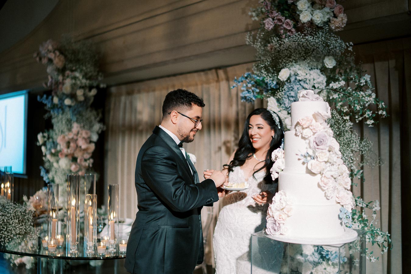 A bride and groom sharing a moment cutting their wedding cake, with a backdrop of elegant floral arrangements. The bride, in a lace wedding dress, smiles as the groom offers her cake. The multi-tiered cake is adorned with pastel flowers, adding to the romantic setting.