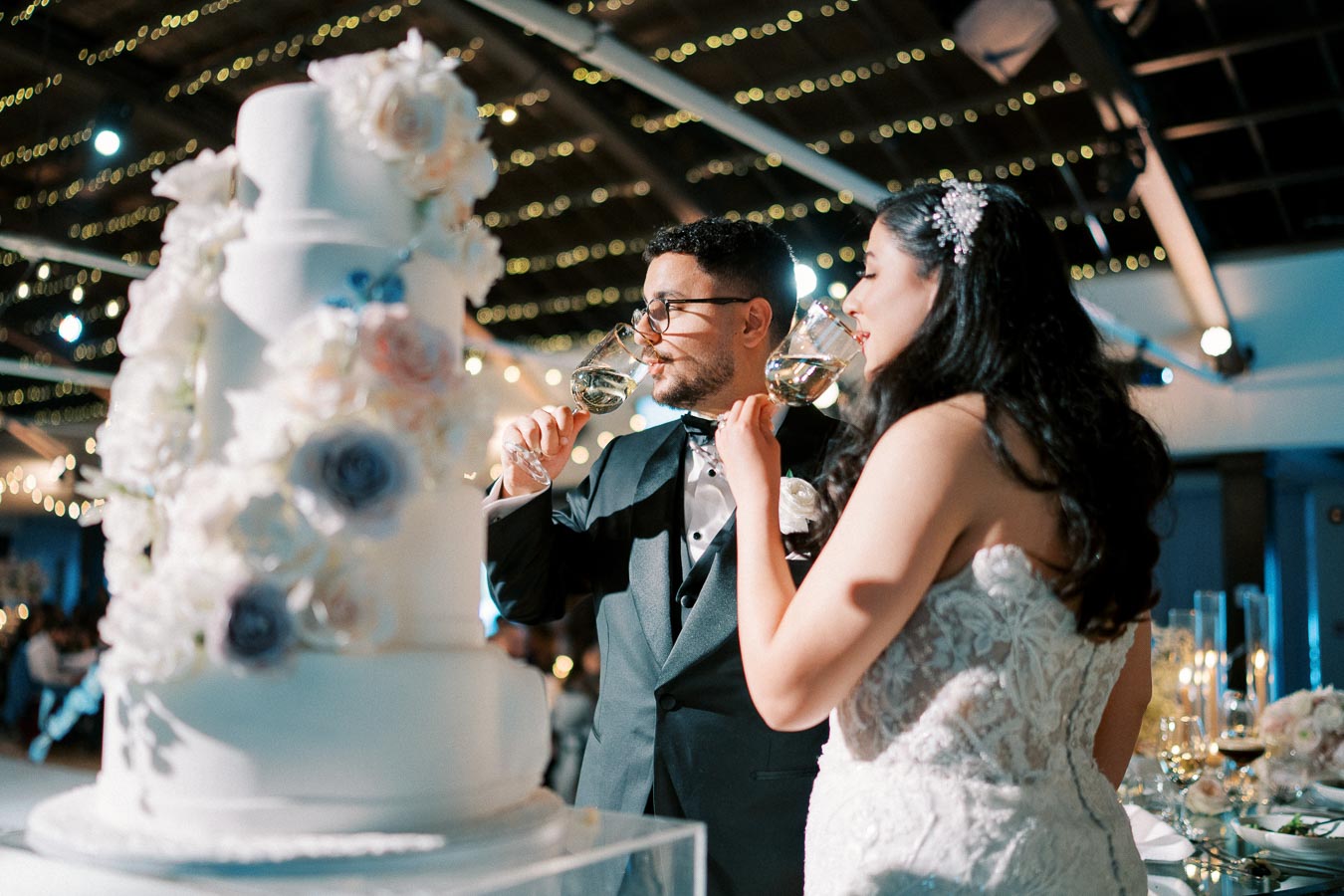Bride and groom toasting with glasses of wine next to a tiered wedding cake adorned with flowers at a beautifully lit reception hall.