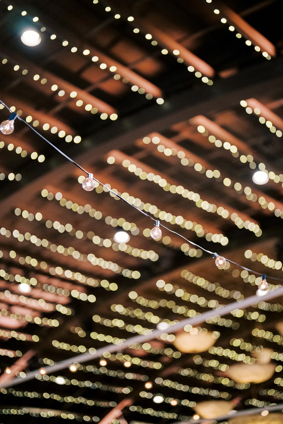 String lights hanging across a rustic wooden ceiling, with warm bokeh effect creating an inviting ambiance. Ideal for event and wedding venue inspirations.