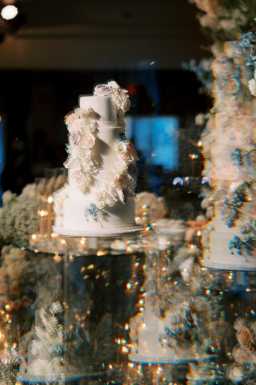 Elegant three-tier white wedding cake adorned with delicate pastel roses, displayed on a glass stand, surrounded by ethereal floral decorations and soft lighting at a sophisticated event.
