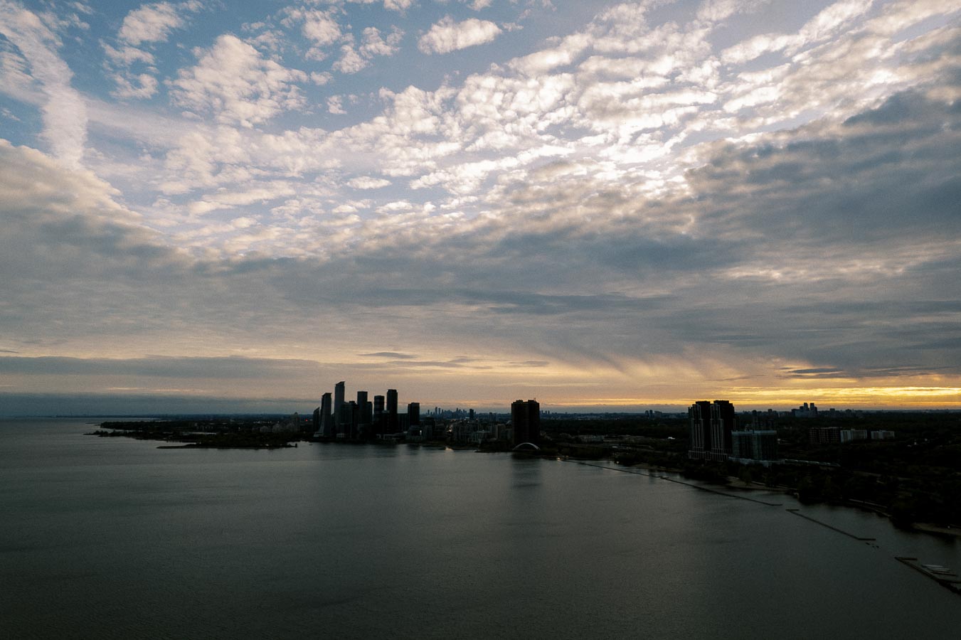 Aerial view of a city skyline against a dramatic cloudy sky at sunset, with tall buildings silhouetted along the waterfront.