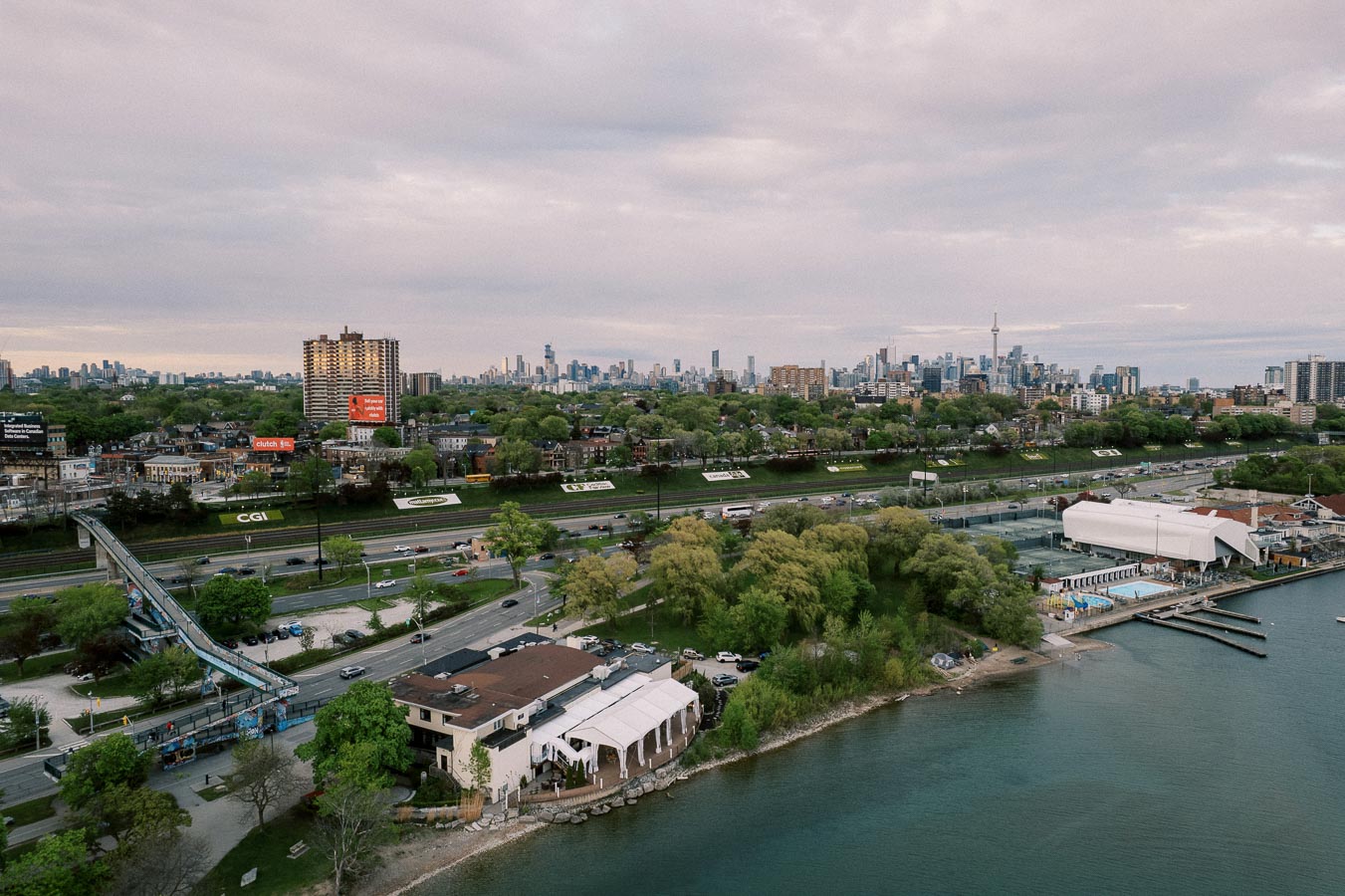 Aerial view of Toronto skyline with lush greenery, Lake Ontario shoreline, and urban landscape under an overcast sky.