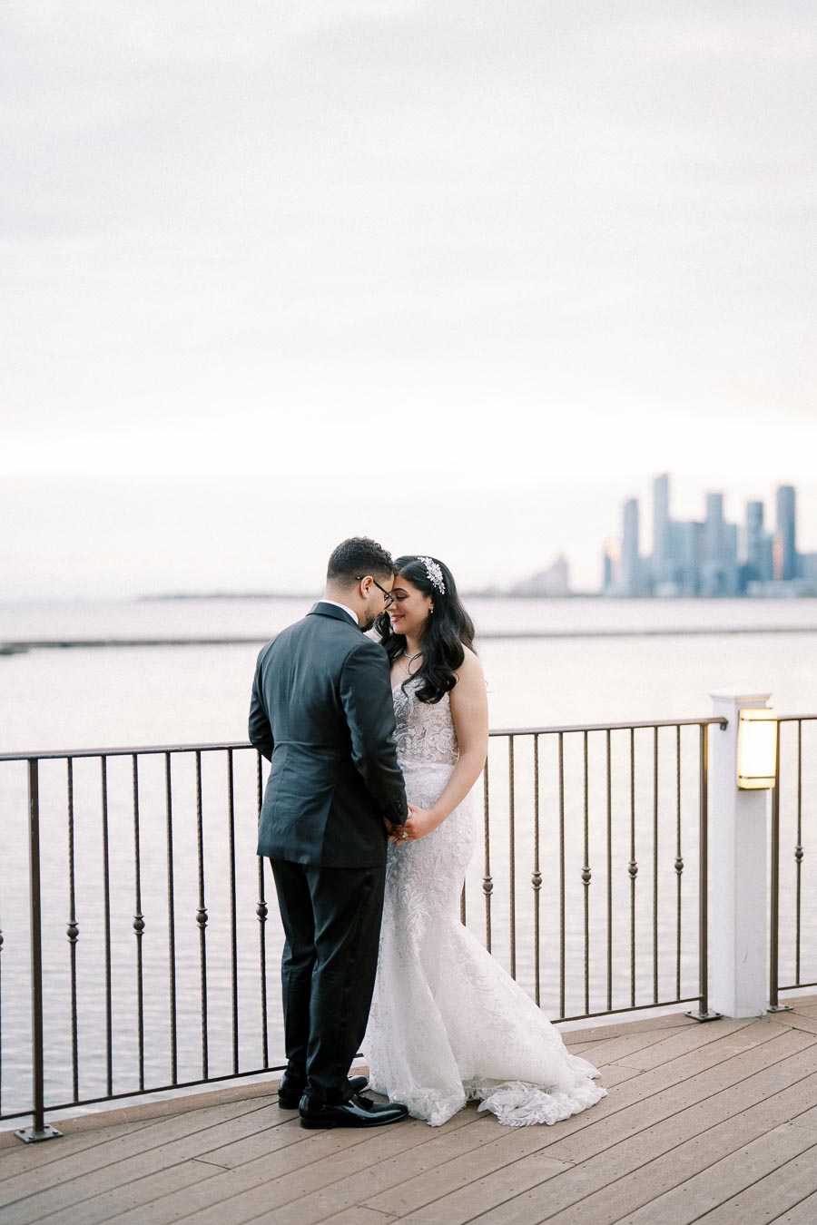 Elegant couple embracing on a waterfront balcony with city skyline backdrop, bride in lace wedding dress and groom in black suit, capturing a romantic wedding moment at sunset.
