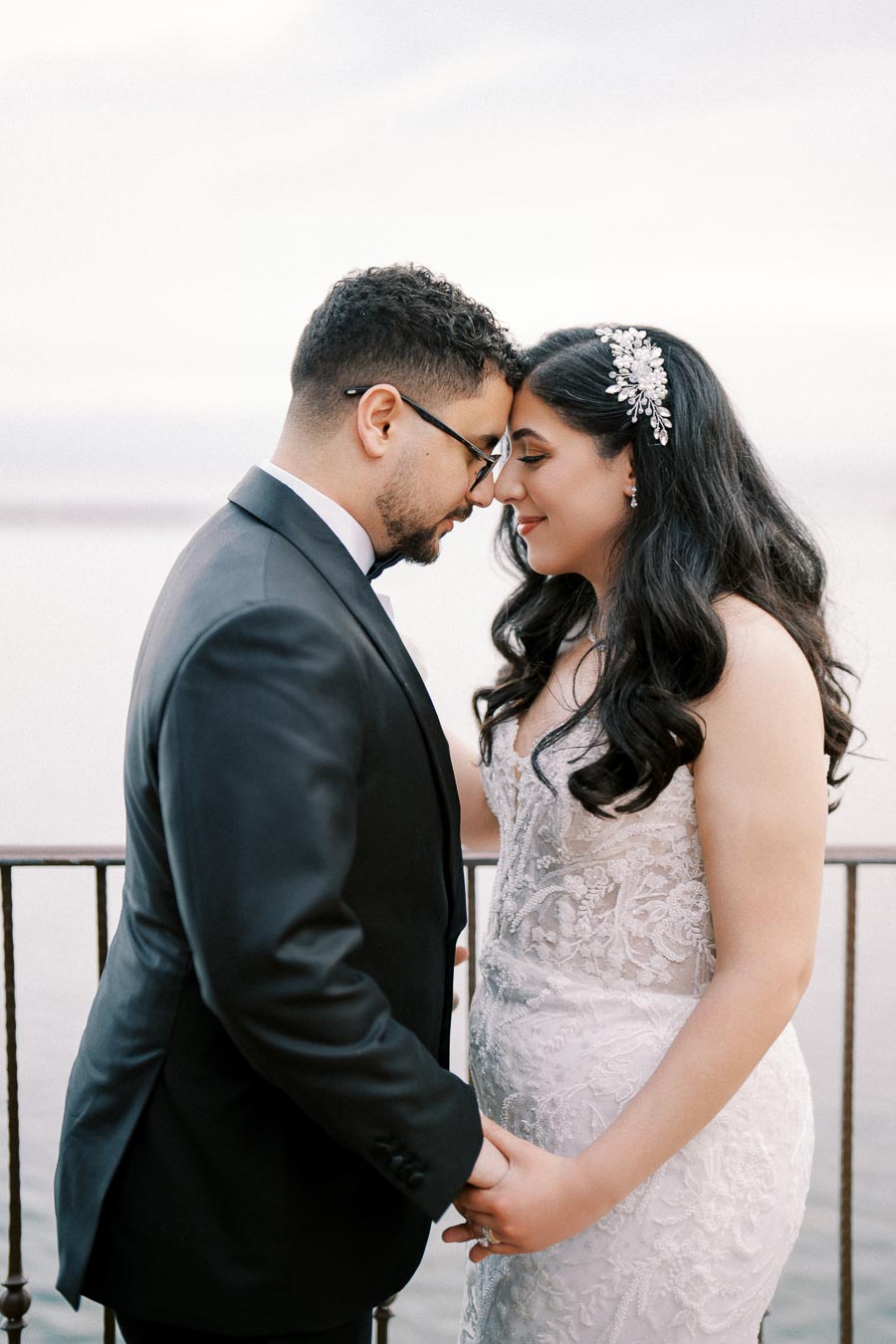 A bride and groom sharing an intimate moment on their wedding day, standing close and holding hands outdoors, with a scenic waterfront backdrop. The bride wears an elegant lace gown and a jeweled hairpiece, while the groom is dressed in a classic black suit and tie.