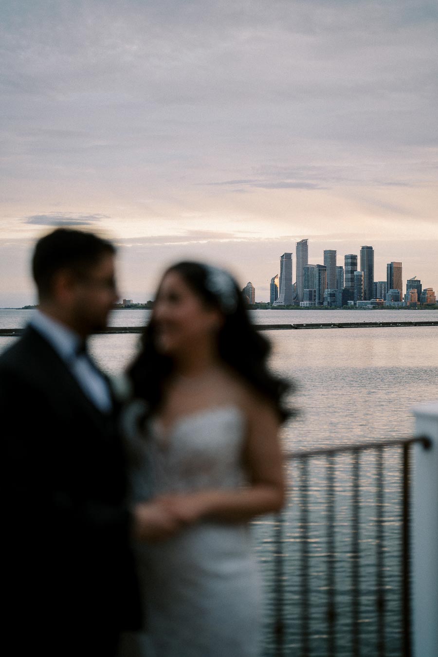 A blurred couple stands in the foreground overlooking a waterfront city skyline at sunset, capturing a romantic and serene atmosphere.