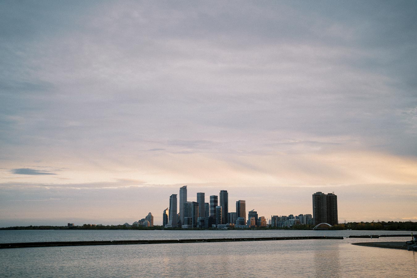 City skyline at dusk with modern skyscrapers reflecting on calm waterfront under a cloudy sky.