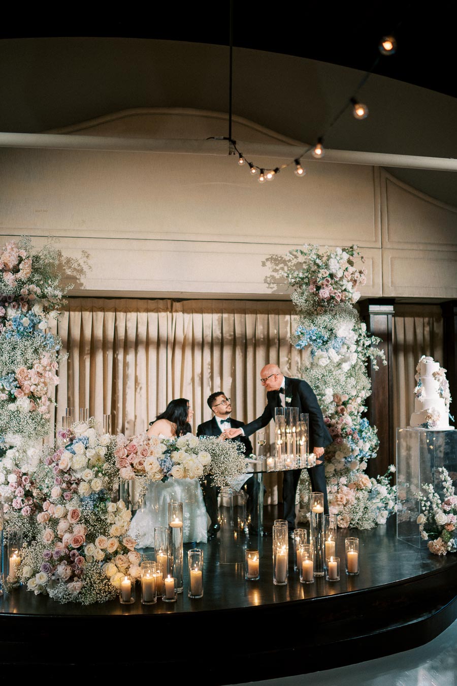 Elegant wedding reception scene with a couple seated at a beautifully decorated table, surrounded by lush floral arrangements and candles. A man in a suit is congratulating them, while a tiered wedding cake stands to the side.
